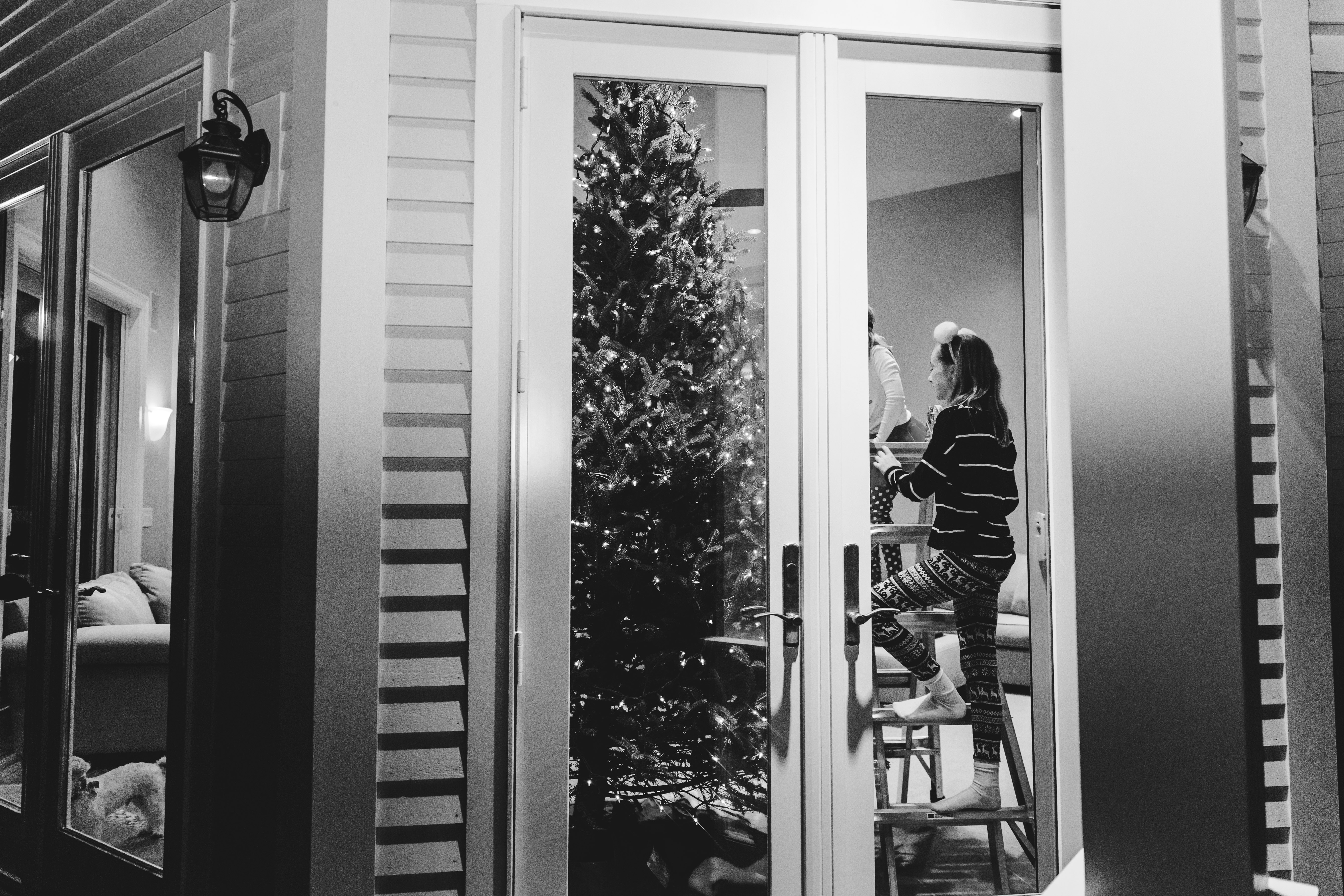 looking in glass doors at girl climbing ladder to decorate Christmas tree