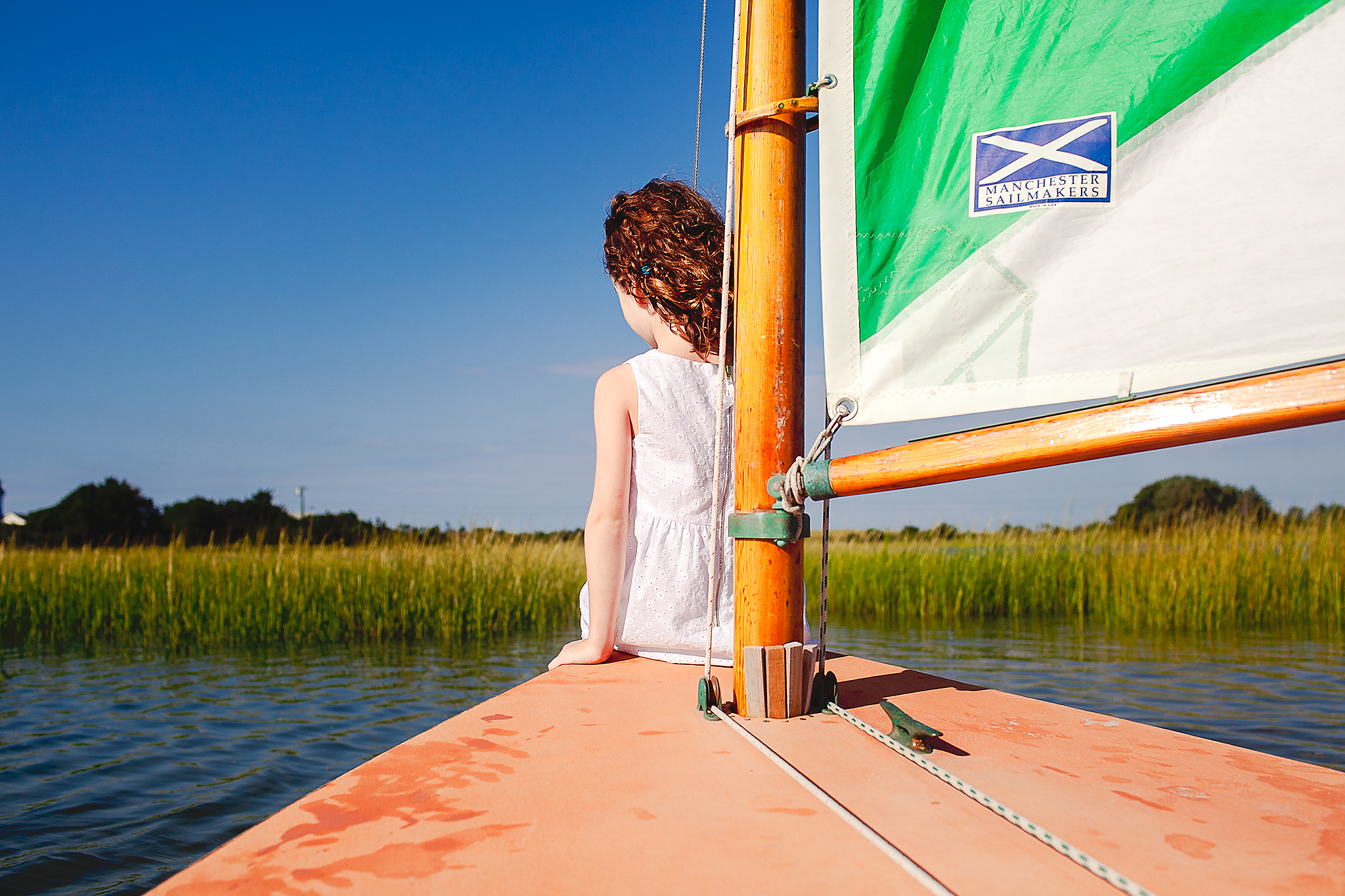 little girl in white dress sitting on front of sailboat heading towards sea grass
