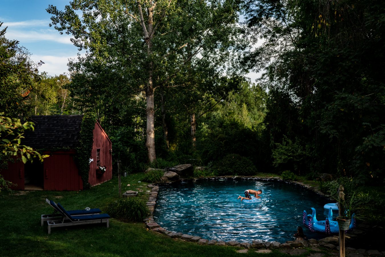 backyard view of pool with boy floating in it and red barn in background