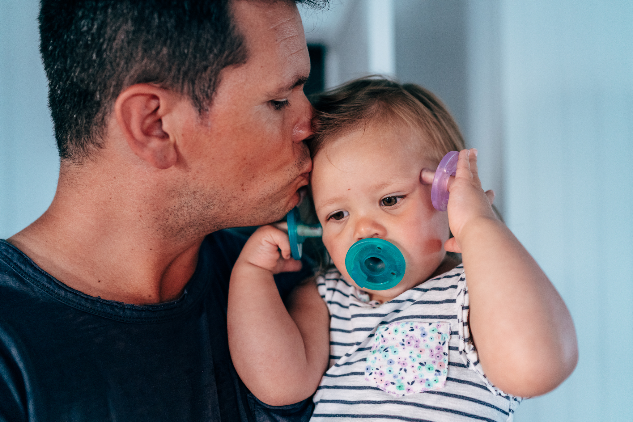 father kissing baby holding pacifiers in hands and mouth
