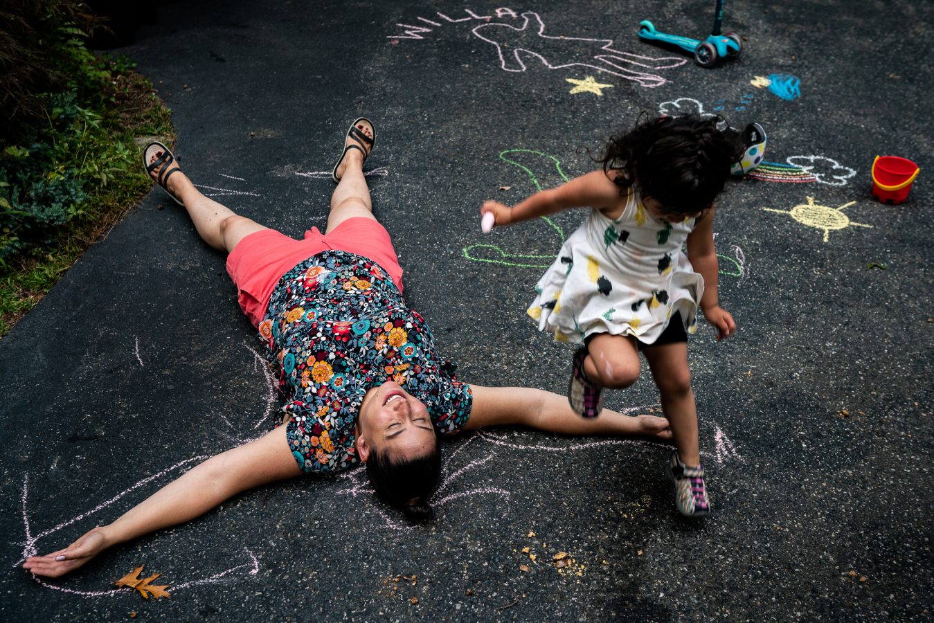 mom lying on driveway with sidewalk chalk drawings as young girl jumps over her arm