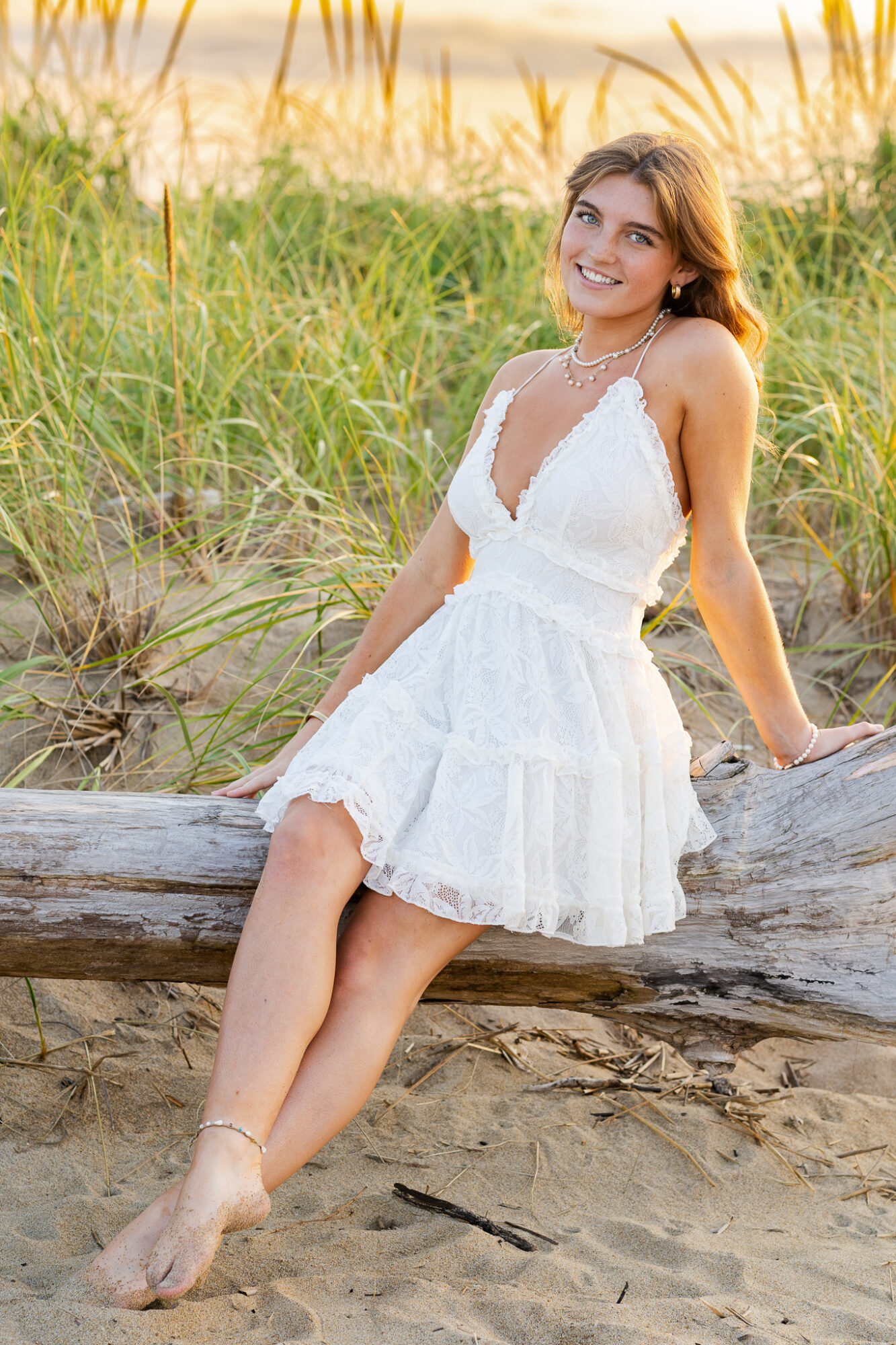 high school senior female wearing white sundress sitting on driftwood at beach