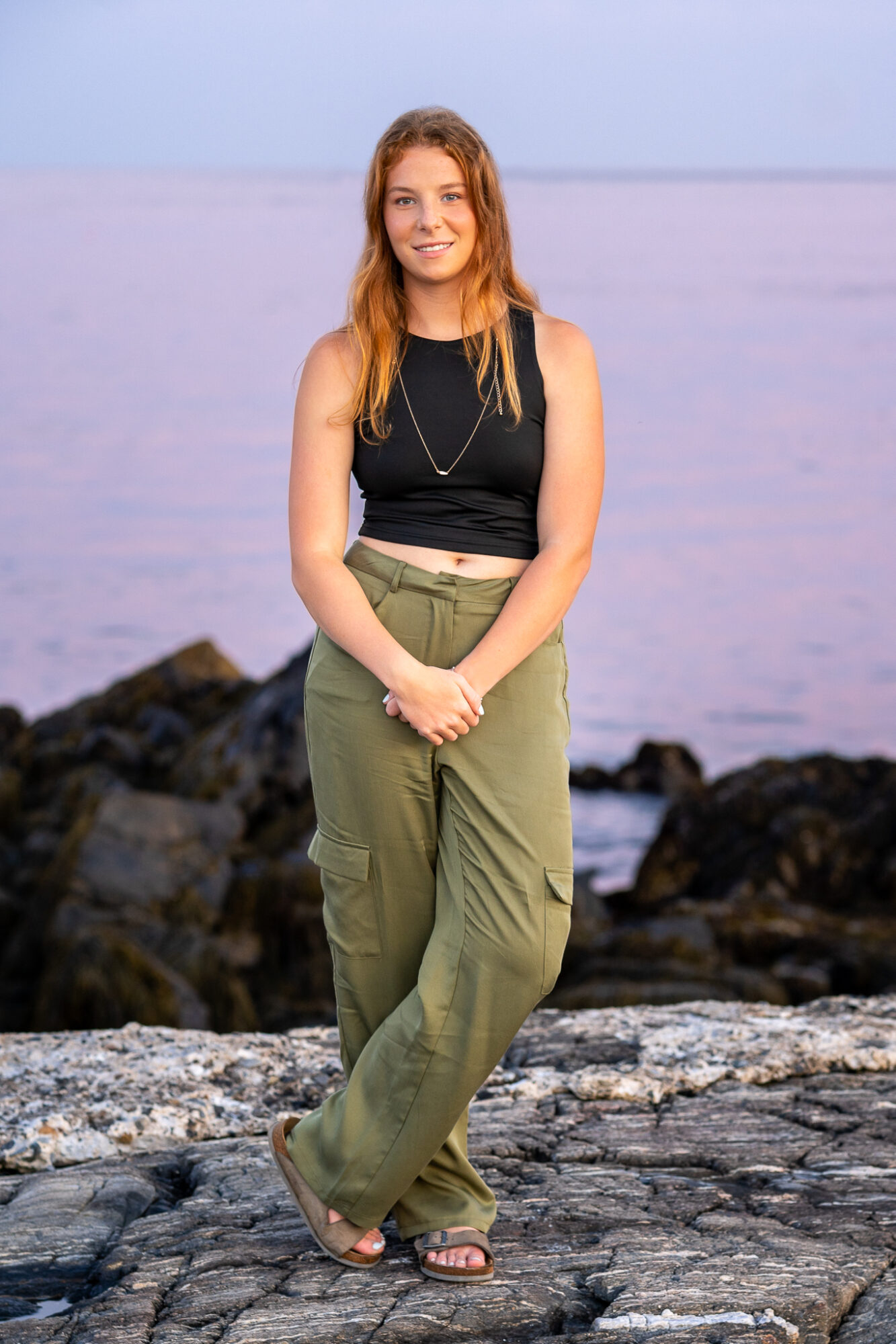 high school senior girl with auburn hair wearing black tank top and green cargo pants standing on rock at sunset