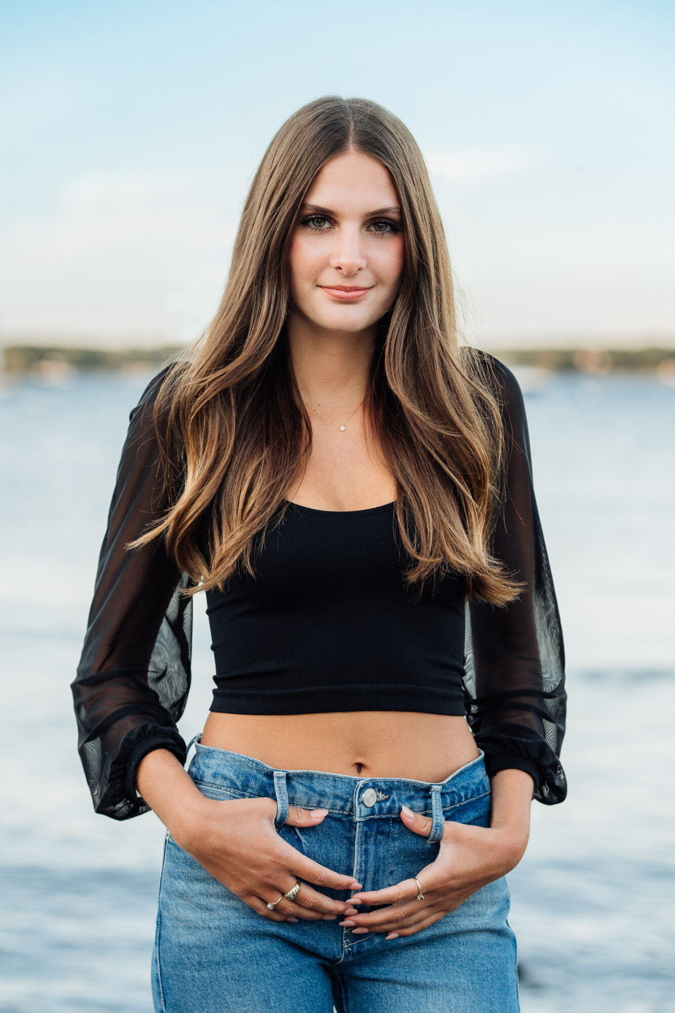 high school senior girl with long brown hair wearing black shirt and jeans by ocean