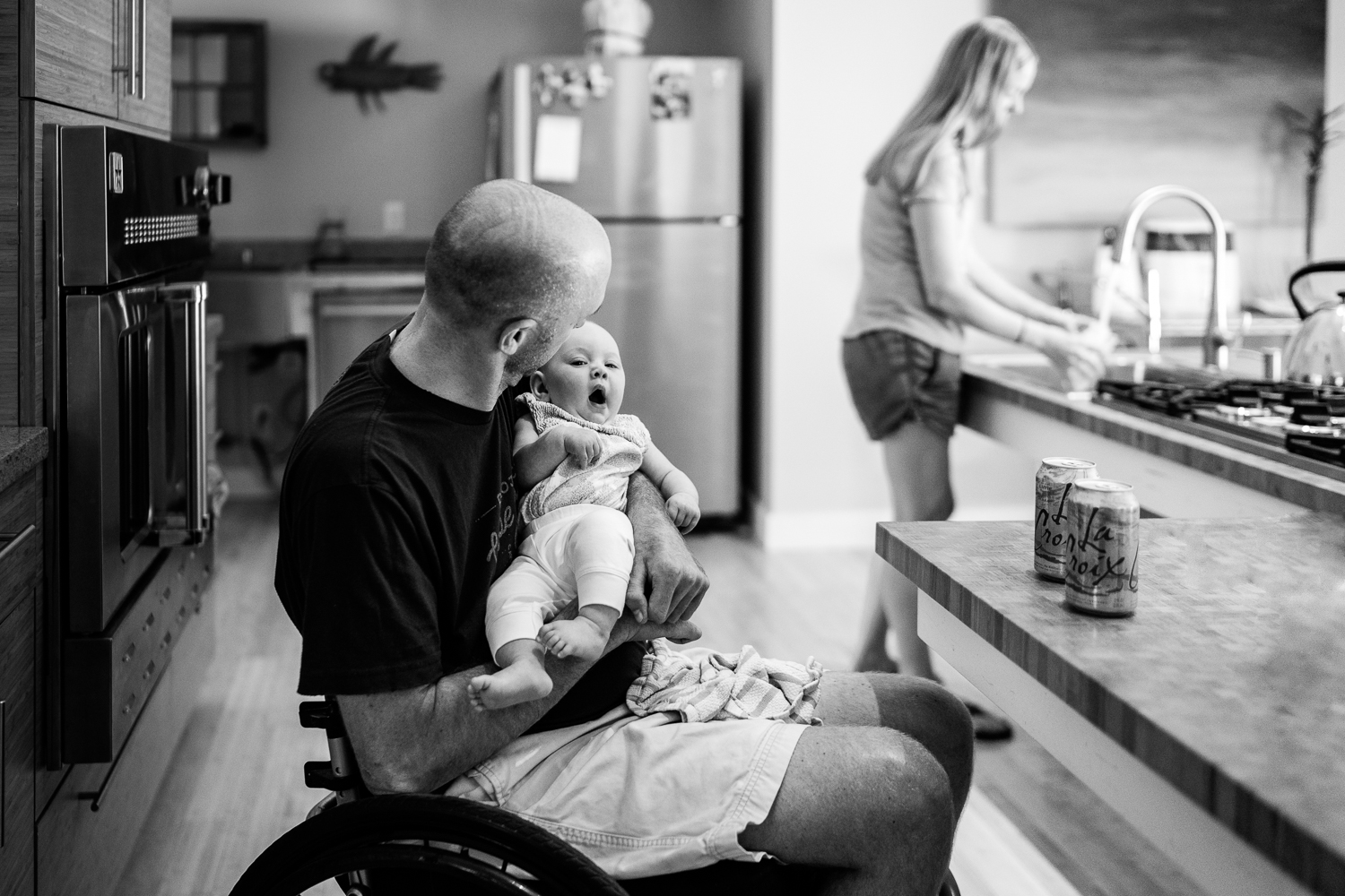 father in wheelchair holding yawning baby while mom washes dishes