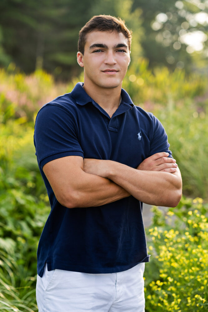 high school senior male wearing navy blue short sleeve collared polo shirt standing with arms crossed in a field