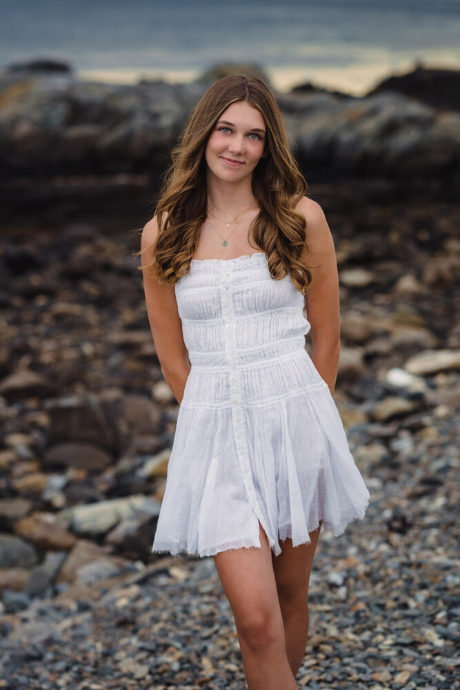 high school senior girl in white dress with long brown hair standing in a rocky area