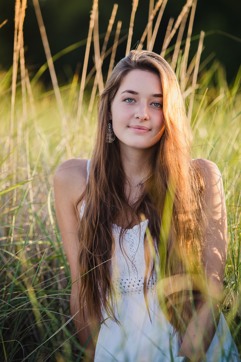 senior portrait of teenage girl sitting in beach grass wearing a white dress
