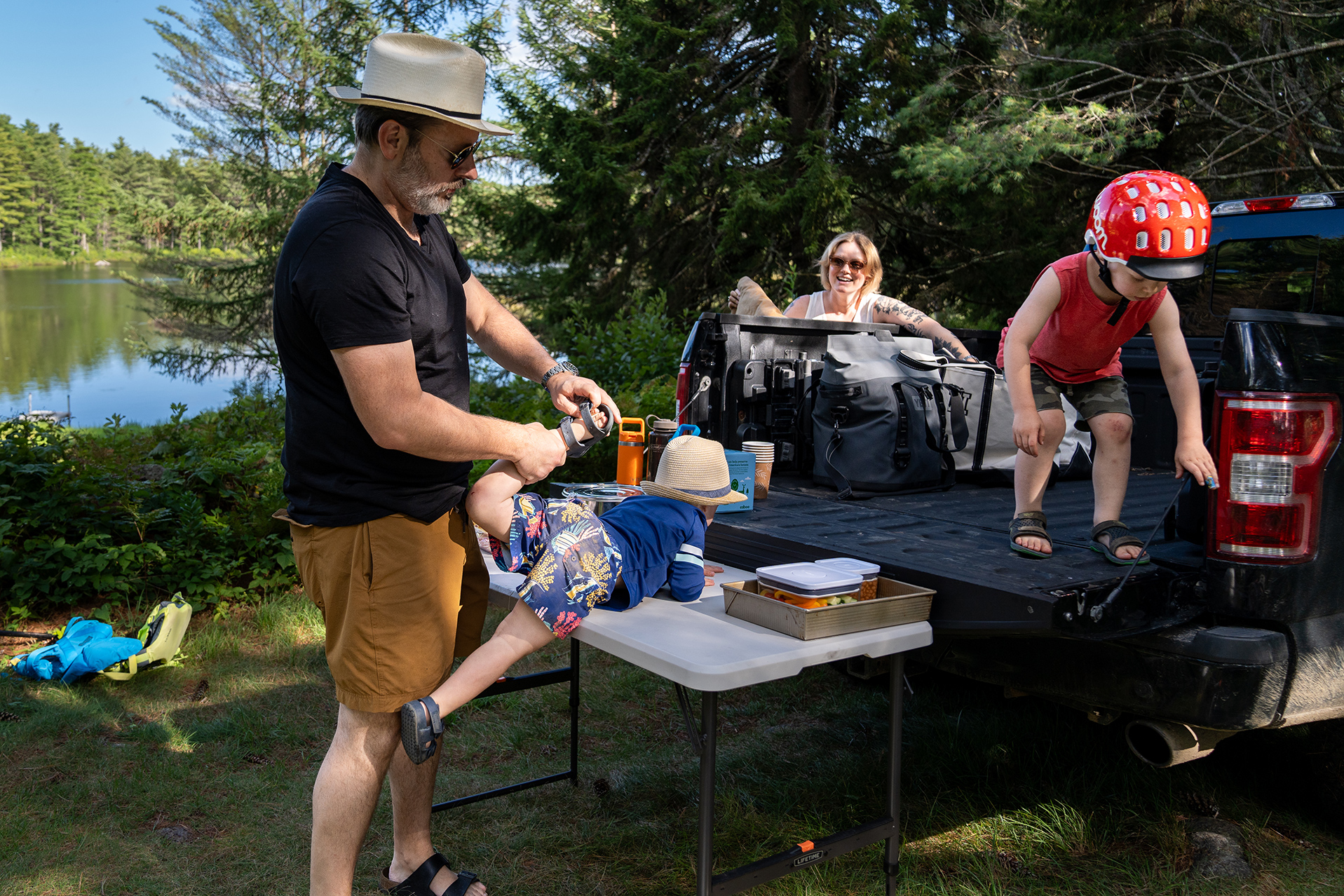 family unpacking a picnic from the back of a pickup truck next to a lake with trees